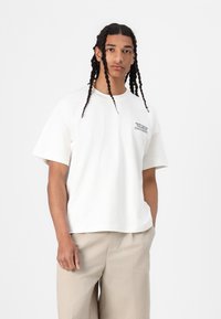 Tricou alb din bumbac, cu mâneci scurte, decolteu rotund și un mic detaliu de text negru pe piept. Modelul poartă pantaloni bej deschis.