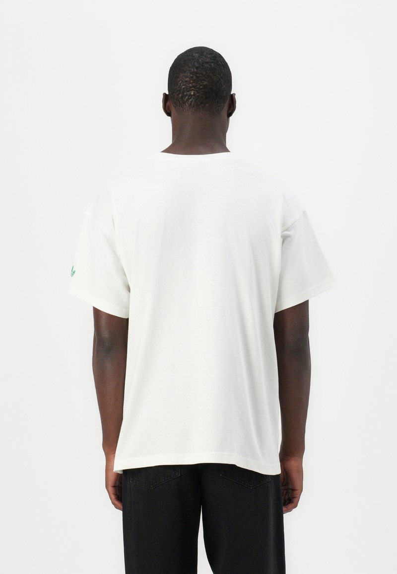Biała bawełniana koszulka o luźnym kroju, z krótkimi rękawami i zielonym akcentem logo na lewym rękawie. Gładka tekstura, minimalistyczny design.
