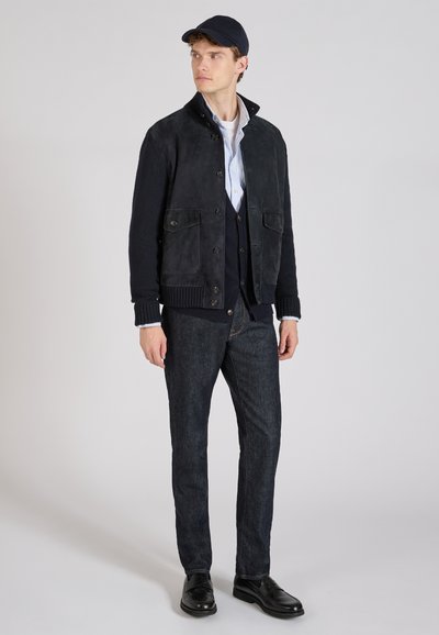 Veste en daim noir avec fermeture à boutons, deux poches avant, manches en tricot, associée à un cardigan bleu marine et un jean sombre, complétée par des chaussures noires.