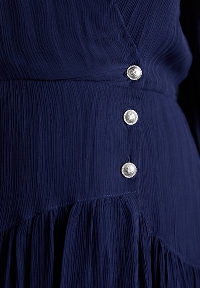 Rochie bleumarin cu un finisaj texturat, având trei butoane decorative argintii pe lateral și o margine cu volane.