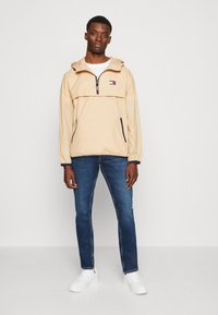 Beige letvægtsjakke med hætte, en brystlomme, navy detaljer og logopræg, parret med blå jeans og hvide sneakers.
