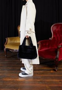 Svart faux fur-väska med två korta handtag, rektangulär form, parat med en beige kappa och svarta sneakers mot en träddäck.