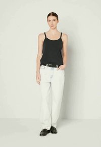Top preto sem mangas, calças largas off-white, cinto preto, sapatos pretos. Modelo está de pé com uma mão no bolso contra um fundo neutro.