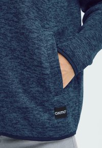 Tumši zils pulovers ar teksturētu rakstu, sānu kabatu un melnu birku pie apakšmalas. Aproces ir tumši zilas ar kontrastu.