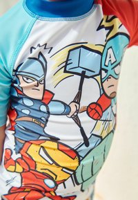 T-shirt med korte ærmer, der har et farverigt comic-inspireret print af superhelte som Thor, Captain America og Iron Man. Farverne er blå, rød og hvid.