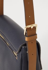 Borsa a spalla in pelle blu navy con una tracolla marrone regolabile e fibbia dorata. Presenta una chiusura con zip e dettagli di cucitura visibili.