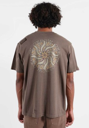 T-shirt en coton marron à manches courtes, avec un grand motif graphique tourbillonnant en blanc et or dans le dos, et une petite étiquette logo sur l'épaule.
