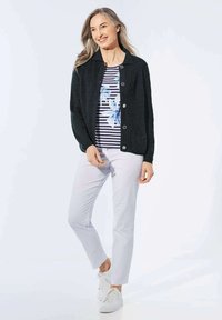 Schwarze Strickjacke mit Kragen, silbernen Knöpfen, kombiniert mit einem gestreiften Oberteil mit blauen Blumenmotiven und weißen Hosen. Weiße Sneakers.