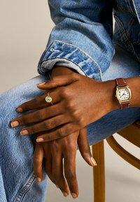 Armbanduhr mit braunem Lederarmband, Gehäuse in Goldton, weißem Zifferblatt und römischen Ziffern. Goldring mit einem zentralen klaren Edelstein an einer Hand, die Hände ruhen auf Denim.
