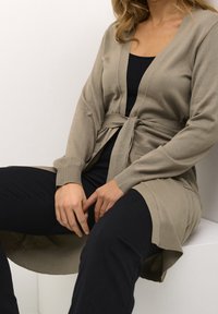 Donna seduta, che indossa un cardigan beige con cintura su una maglietta nera e pantaloni neri, con le mani appoggiate sulle ginocchia su uno sfondo bianco semplice.