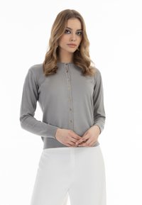 Grauer Langarm-Cardigan mit goldenen Knöpfen, gerippten Bündchen und Saum sowie einer taillierten Passform. Über weißen Hosen getragen. Glatte Textur.