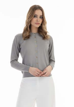 Cardigan grigio a maniche lunghe con bottoni dorati, polsini e orlo a coste, e un design aderente. Indossato sopra pantaloni bianchi. Texture liscia.