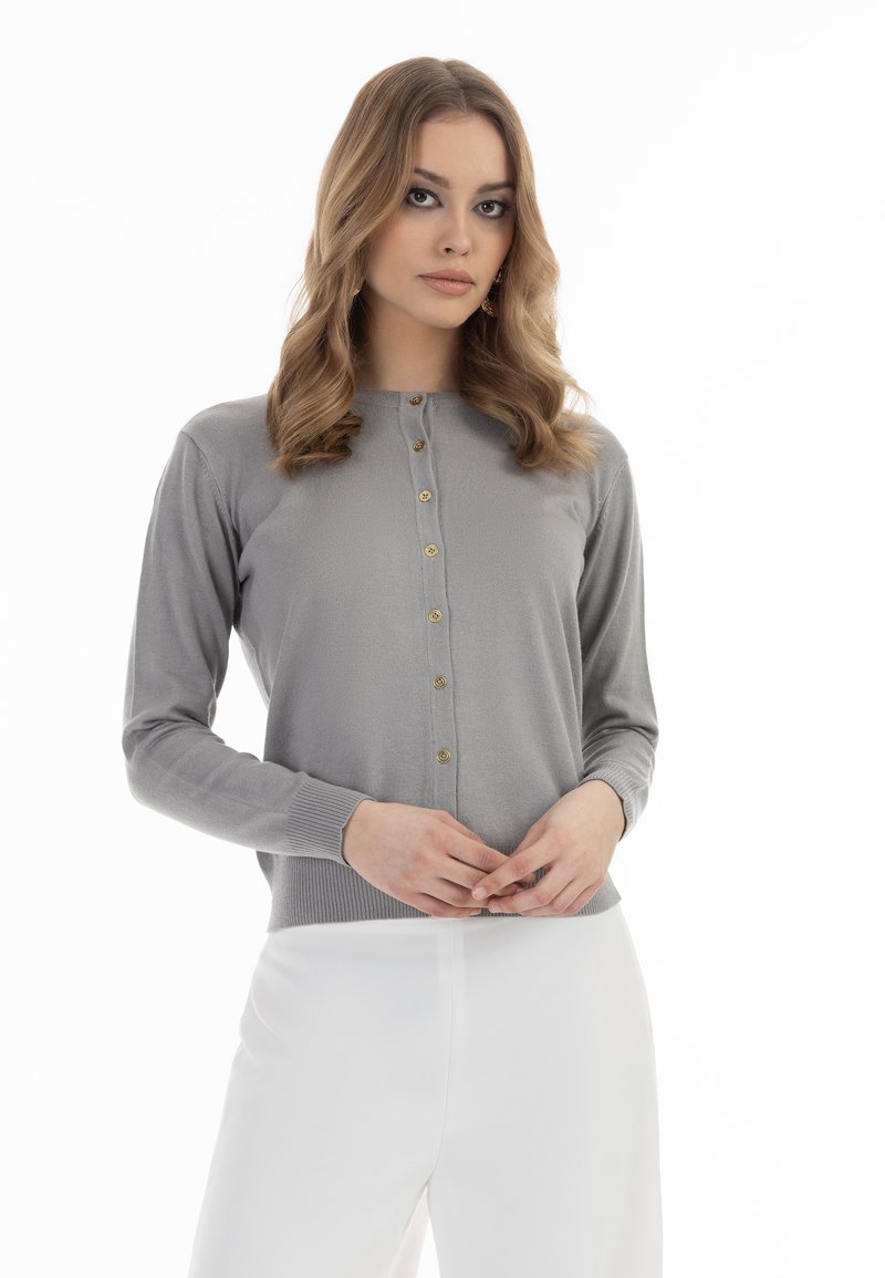Grauer Langarm-Cardigan mit goldenen Knöpfen, gerippten Bündchen und Saum sowie einer taillierten Passform. Über weißen Hosen getragen. Glatte Textur.