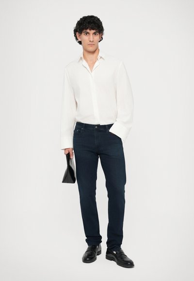 Λευκό πουκάμισο με μακριά μανίκια, σκούρες μπλε εφαρμοστές τζιν και μαύρα παπούτσια. Κρατάει μια μικρή μαύρη τσάντα. Απλό φόντο, χωρίς σχέδια ή υφές.