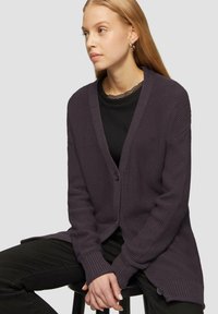Lila gestrickte Strickjacke aus strukturiertem Stoff, mit offenem Vorderteil, langen Ärmeln und Knopfdetails am Saum. Über einem schwarzen Oberteil getragen.