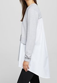 Una donna indossa un top a strati con una felpa grigia davanti e un tessuto simile a una camicia bianca che si estende sul retro, abbinato a pantaloni neri.