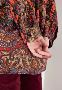 Colorful patterned shirt featuring floral designs in orange, pink, and black, with a decorative cuff and buttons, paired with dark red pants.