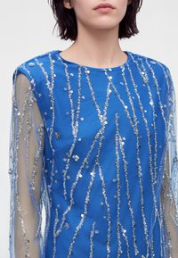 Femme portant un haut bleu transparent à manches longues décoré de sequins argentés verticaux et d'ornements floraux.