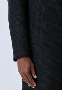 Tmavé kárované vlnené sako so vzpriameným, štruktúrovaným dizajnom. Obsahuje jednu bočnú vrecko a prispôsobený rukáv s preložením na manžete.