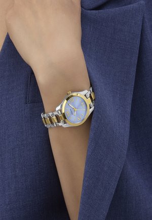 Silver and gold-tone metal watch with a round blue dial, Roman numerals, and a linked bracelet design. Worn on a wrist near a blue fabric.