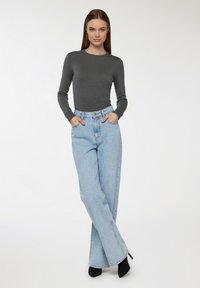 Szary długi rękaw bluzka w zestawieniu z jasno-niebieskimi spodniami typu high-waist o szerokich nogawkach, widoczne kieszenie z przodu, noszone z czarnymi szpilkami, na jednolitym tle.