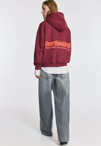 Hanorac bordo cu text portocaliu pe spate, asortat cu blugi gri largi. Designul include o glugă și manșete cu refiletat, având un aspect stratificat.