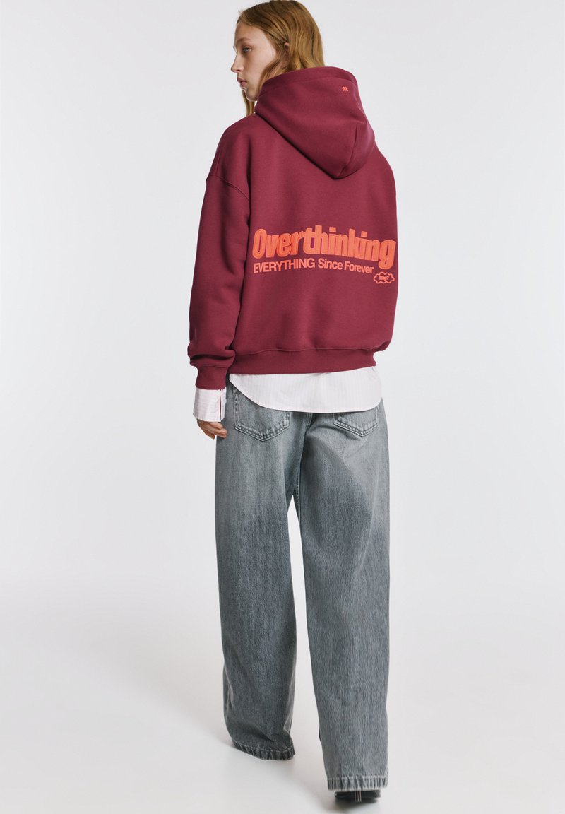 Hanorac bordo cu text portocaliu pe spate, asortat cu blugi gri largi. Designul include o glugă și manșete cu refiletat, având un aspect stratificat.