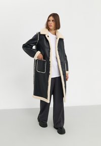 Cappotto in pelle nera con colletto e rifiniture in shearling crema, dotato di chiusura a bottoni e tasca frontale a inserto. Design lungo e dal fit morbido.