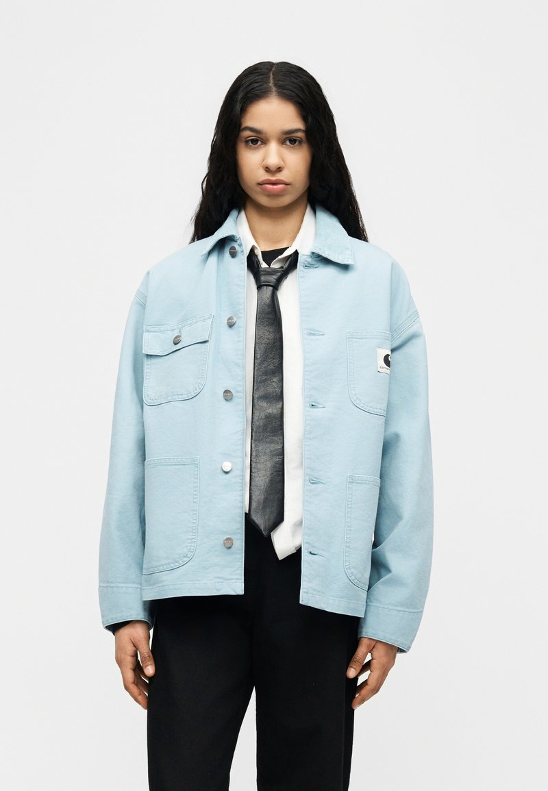 Woman with long dark hair wearing light blue jacket, white shirt, black tie, and black pants standing against plain background.