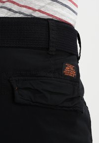 Pantaloni cargo neri in tessuto con cintura elastica texturizzata. Presenta una tasca a toppa sulla coscia e un'etichetta con accenti arancioni.