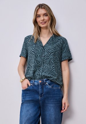 Vrouw draagt een donkergroen zebraprint kort mouw overhemd met knopen, ingestopt in een blauwe high-waisted spijkerbroek, met een gouden armband om haar linkerpols.