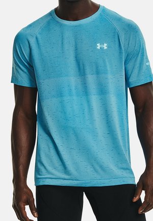 Mann trägt enganliegendes hellblaues Under Armour kurzärmeliges Sportshirt und schwarze Hose, steht mit entspannt an den Seiten hängenden Armen.