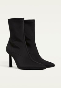 Čierne členkové topánky vyrobené z hladkej textílie, s špicatými prstami a štíhlými vysokými podpätkami. Bezšvový dizajn s jemnými švovými detailmi.