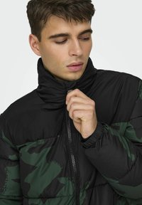 Giubbotto imbottito camouflage nero e verde con colletto alto, zip frontale e polsini elastici. Il materiale ha una finitura liscia e lucida.