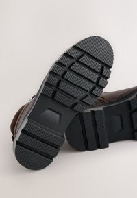 Braune Leder-Stiefeletten mit einer strukturierten schwarzen Gummisohle, die tiefe Profilmuster und ein rundes Zehendesign aufweist.