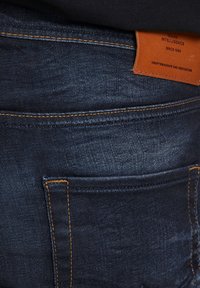 Pantalones vaqueros de mezclilla azul oscuro con costuras naranjas, que cuentan con una etiqueta de cuero en la parte trasera, destacando la artesanía y la dedicación.