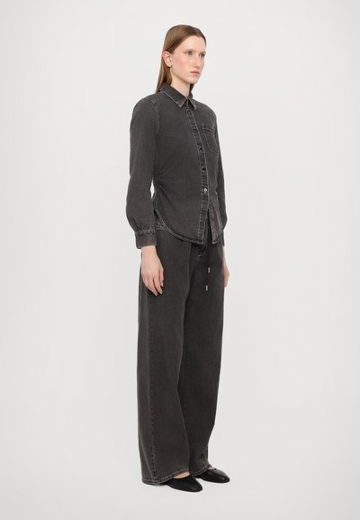 Camisa de ganga preta com botões, ligeiro brilho e bolso no peito; combinada com calças de ganga preta de perna larga. Design simples, sem padrões visíveis.