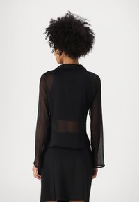 Blouse noire transparente avec des manches longues et un col. Texture douce avec une coupe décontractée, associée à une jupe noire pour le contraste.