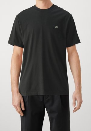 Mężczyzna ubrany w prosty czarny t-shirt z krótkim rękawem z małym zielonym logo krokodyla na lewej piersi, w zestawieniu z czarnymi spodniami.