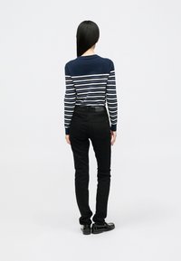 Mulher com cabelo preto longo, vestindo um suéter listrado azul-marinho e branco, jeans pretos e mocassins pretos, de pé de costas em fundo simples.