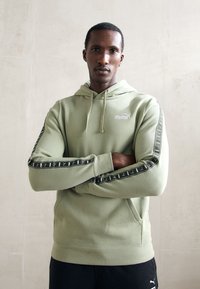 Mand med armene korslagt iført en lys grøn Puma hoodie med sort logo tape på ærmerne og sorte bukser, stående imod en ensfarvet væg.