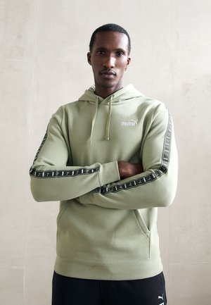 Vīrietis ar rokām šķērsām, ģērbies gaiši zaļā Puma kapučos ar melnu logotipa lente uz piedurknēm un melnām biksēm, stāv pretī vienkāršai siena.