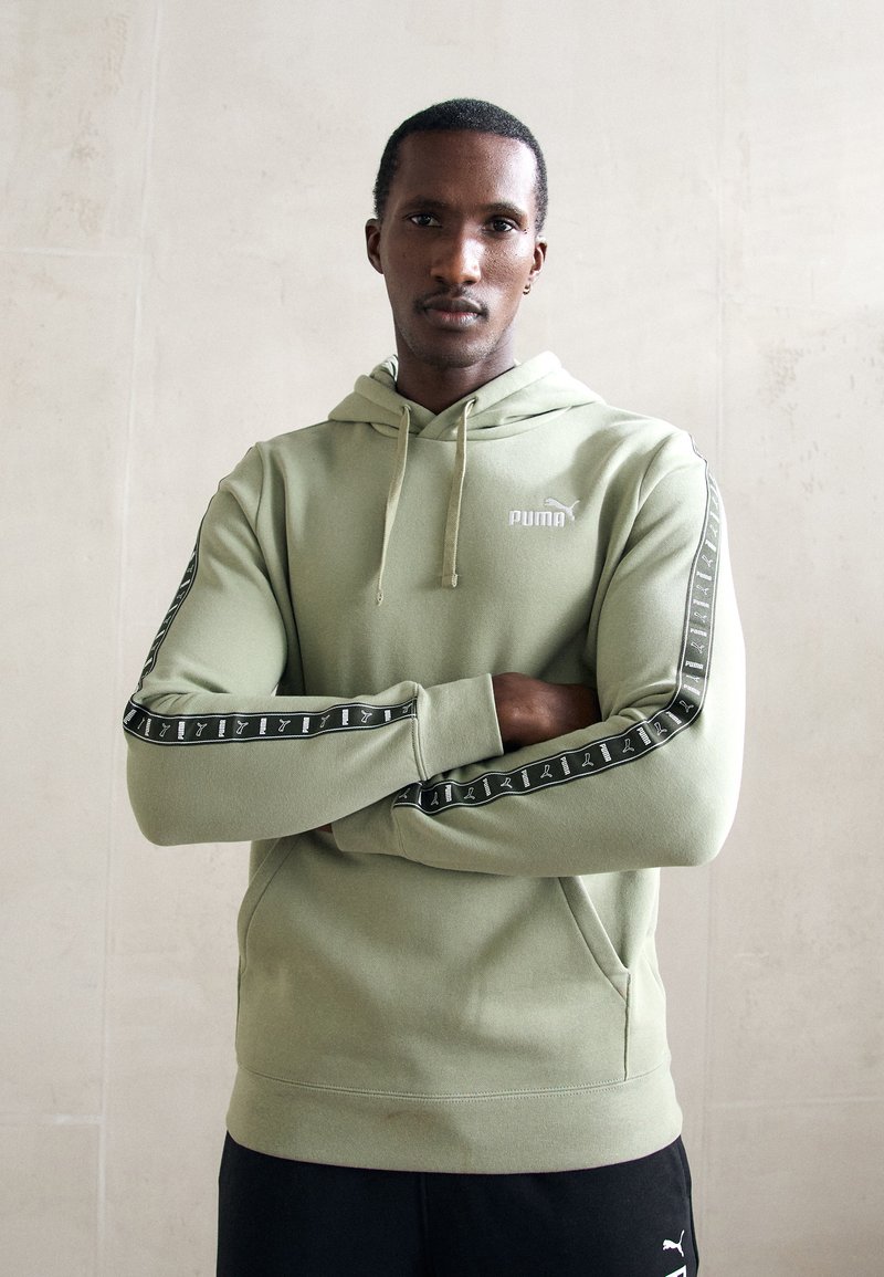 Uomo con le braccia incrociate che indossa un hoodie Puma verde chiaro con nastro logo nero sulle maniche e pantaloni neri, in piedi di fronte a un muro semplice.