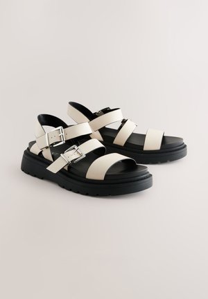 Sandales en cuir blanc avec semelle plateforme noire, comportant plusieurs lanières avec boucles ajustables. Texture lisse, design à bout ouvert.