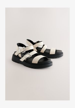 Sandales en cuir blanc avec semelle plateforme noire, comportant plusieurs lanières avec boucles ajustables. Texture lisse, design à bout ouvert.