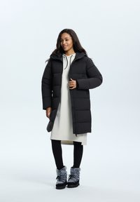 Zwarte pufferjas met een capuchon, ritsclosure en zijzakken, gelaagd over een lichtgrijze jurk, gecombineerd met zwarte leggings en fluffy boots.