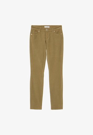 Femme portant un pull à col roulé rose vif, un pantalon en velours côtelé marron clair et des mocassins noirs épais, debout contre un mur blanc.