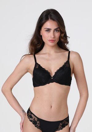 Donna che indossa un reggiseno e slip in pizzo nero coordinati, in piedi con una mano sul fianco contro uno sfondo chiaro e uniforme.