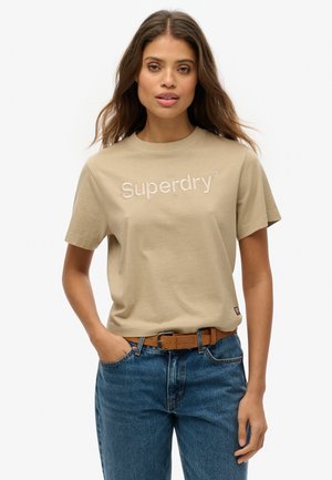 Camiseta de algodón color beige con mangas cortas, que presenta el logotipo "Superdry" bordado en un tono más claro. Combinada con jeans azules y un cinturón marrón.