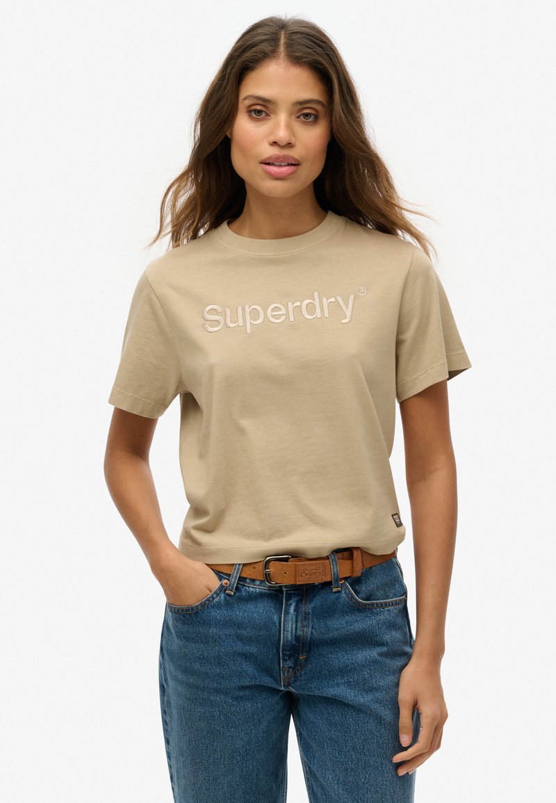 Beige katoenen t-shirt met korte mouwen, voorzien van geborduurd "Superdry" logo in een lichtere schaduw. Gecombineerd met blauwe jeans en een bruine riem.